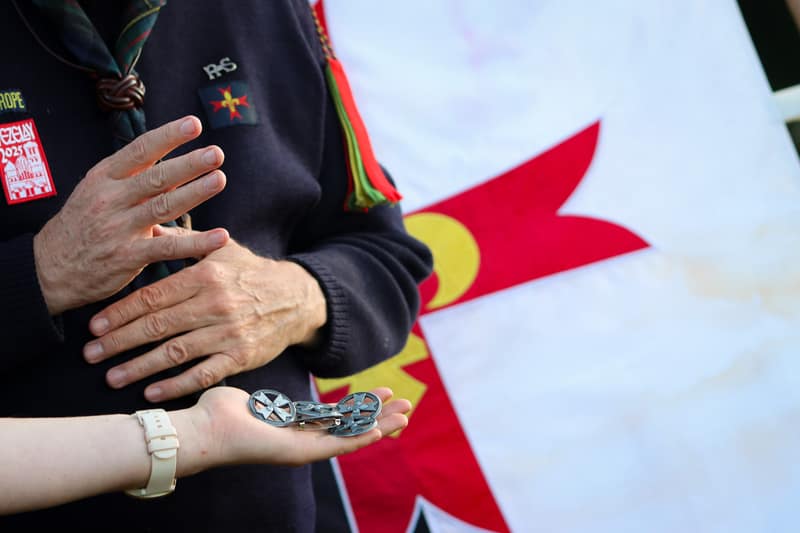 Een jonge hand reikt een scoutsinsigne aan, voor de vlag van de beweging.